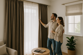 Couple adjusts taupe blackout curtains beside a beige Roman blind and sheer drapes in a sunlit living room, with fabric swatches on the table - affordable luxury window dressing.